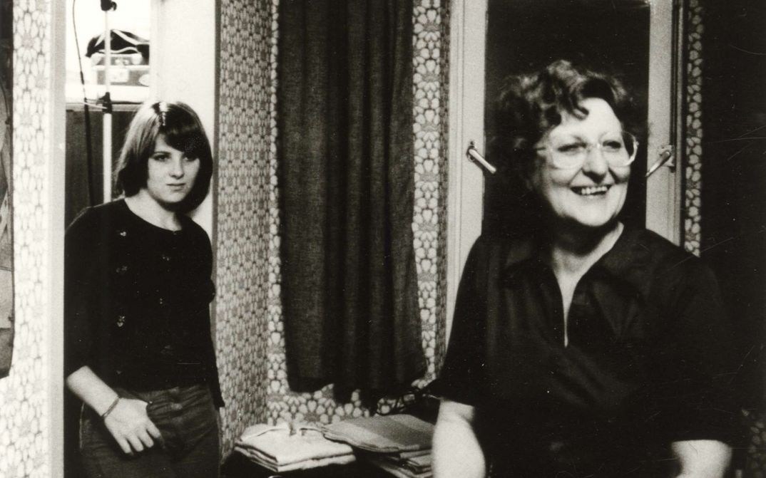 Film still from VON WEGEN SCHICKSAL: A woman stands laughing in the living room, a teenager leans against the doorframe.