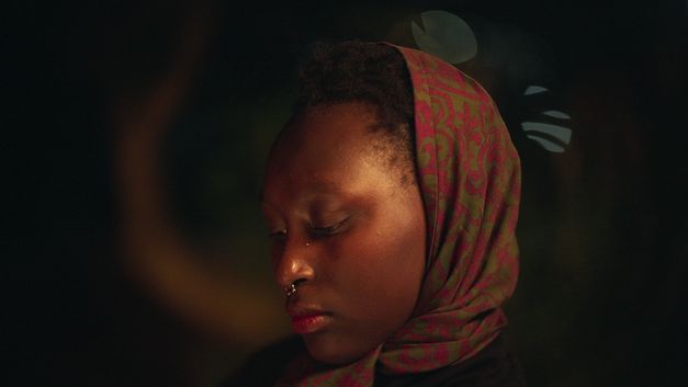 A close-up of a young woman wearing a green and red patterned headscarf against a dark background. 