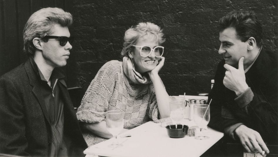 Filmstill aus FRÄULEIN BERLIN: Zwei Männer und eine Faru sitzen an einem Tisch. Sie haben Getränke, tragen große Brillen und rauchen teils.