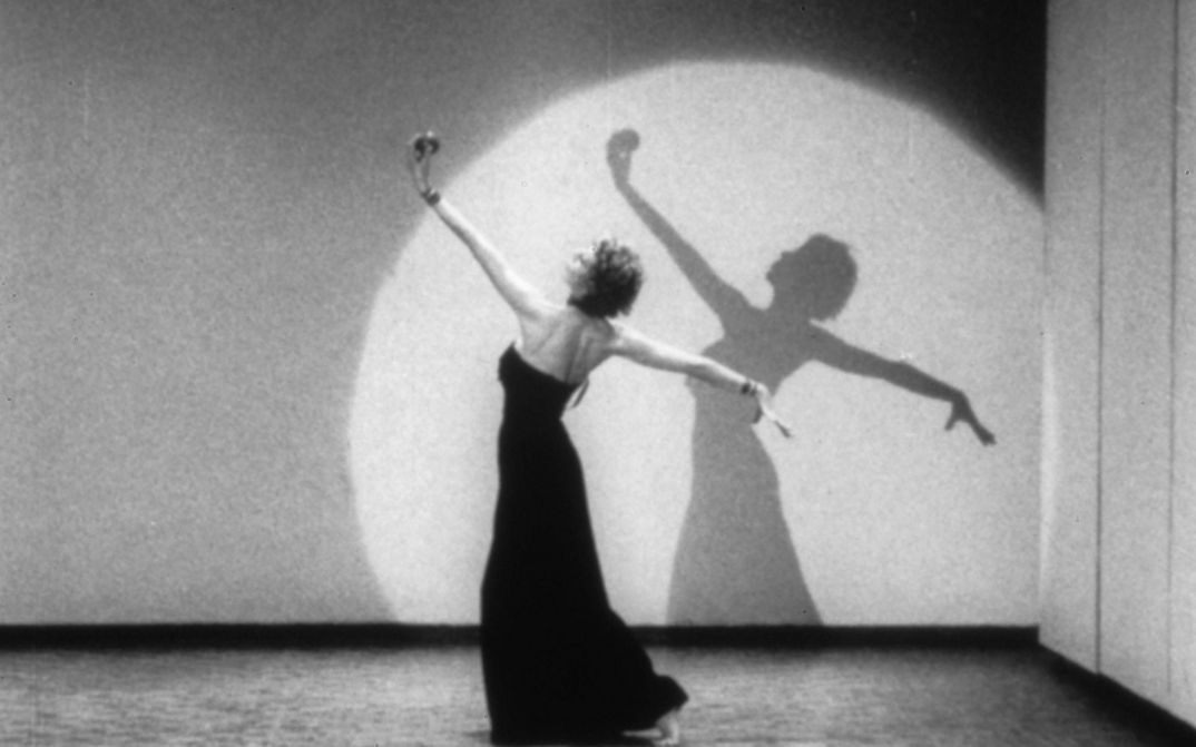 Filmstill aus LIVES OF PERFORMERS: Eine tanzende Frau in einem langen Kleid in einem Lichtkegel.