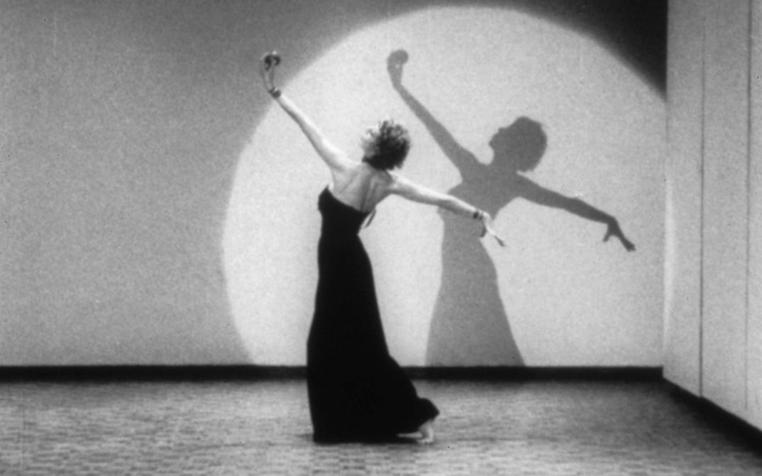 Film still from LIVES OF PERFORMERS: A dancing woman in a long dress in a cone of light.