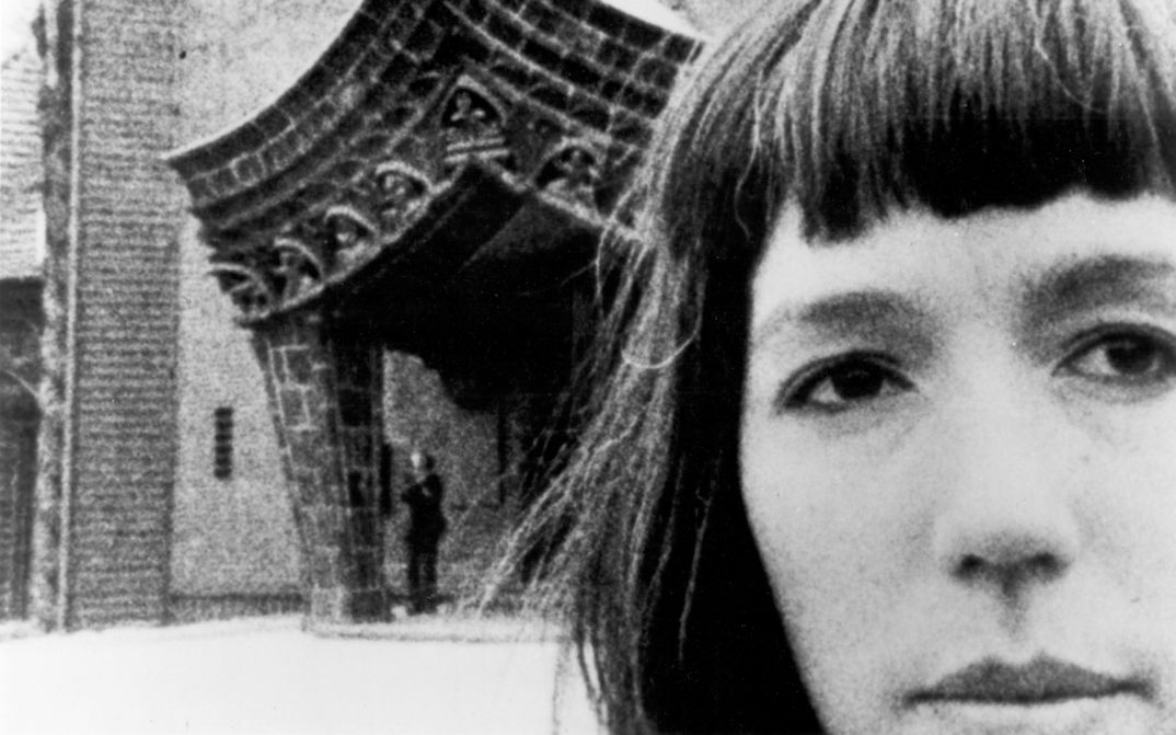 Filmstill aus JOURNEYS FROM BERLIN/1971: Ein Frauengesicht in Großaufnahme, dahinter ein auffällig gebautes Tor.