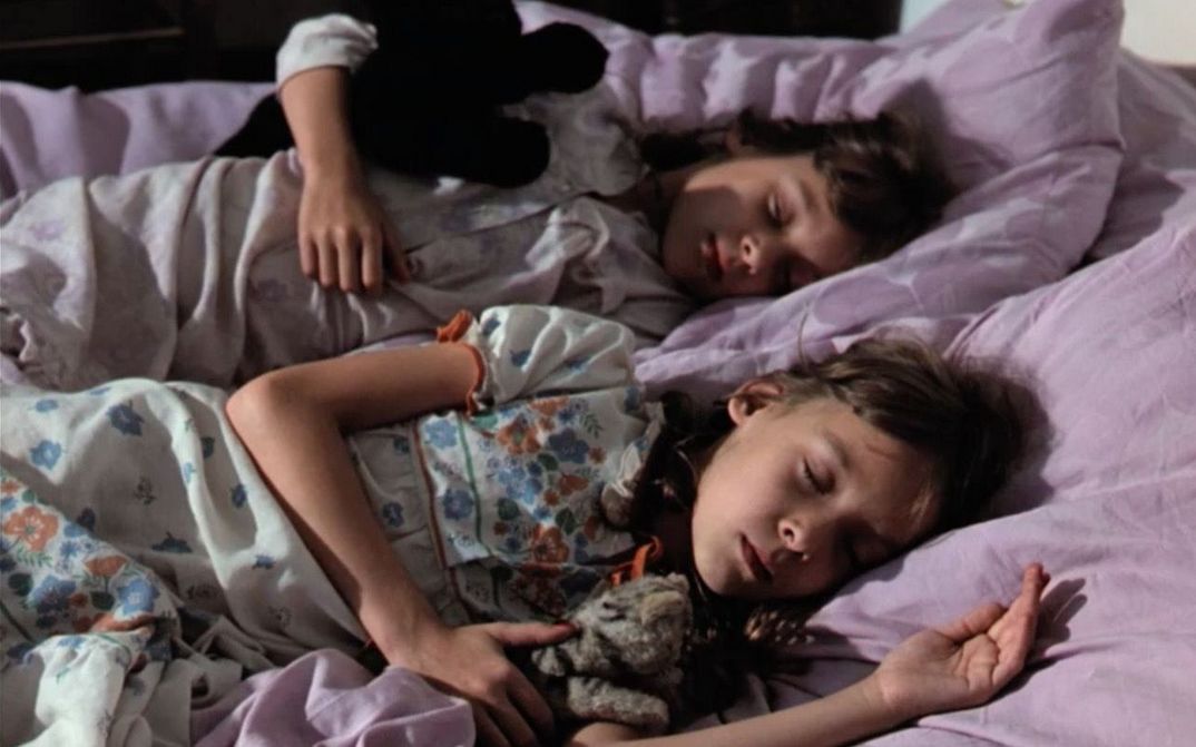 Film still from STAUNEN: two young girls sitting lying next to each other in bed, asleep. One is holding a cuddle toy, the sheets are light purple.