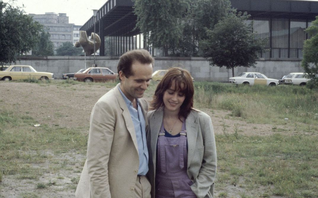 Film still from BERLIN CHAMISSOPLATZ: A man and a woman stand close together on a wasteland in front of the Neue Nationalgalerie in Berlin.