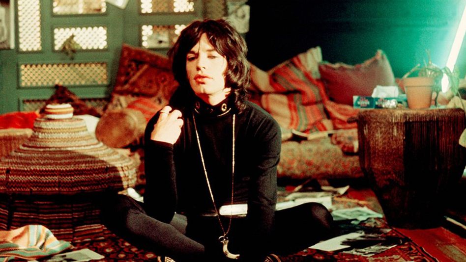 Filmstill aus PERFORMANCE: Mick Jagger sitzt schwarz gekleidet im Schneidersitz auf dem Boden eines Zimmers, das mit farbigen Möbeln und Teppichen ausgestattet ist.