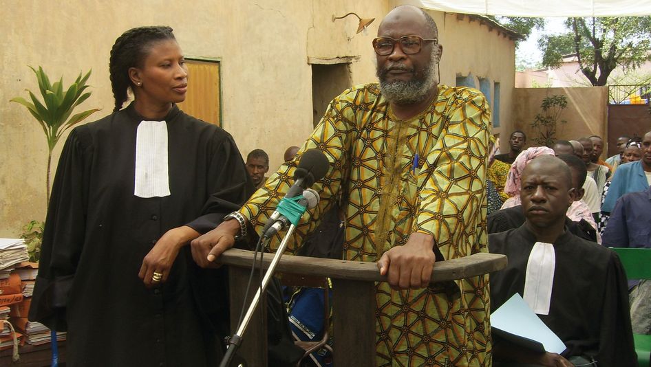 Film still from BAMAKO: An improvised courtroom scene in the open air. In the foreground a woman in a judge's robe, next to her a man is making a statement.