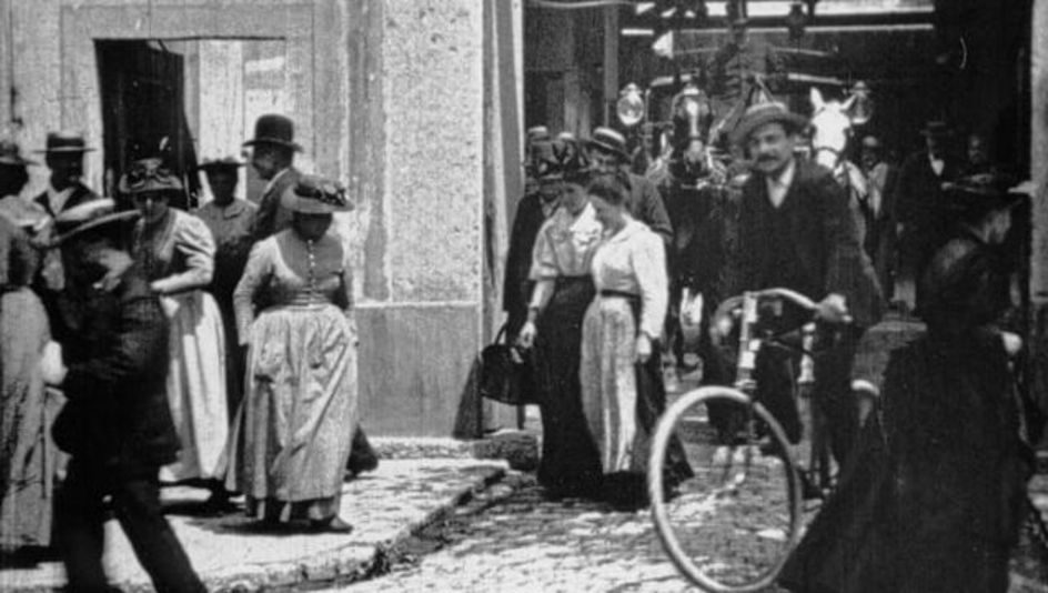 Filmstill aus ARBEITER VERLASSEN DIE FABRIK: Eine Straßenszene; viele Menschen in alten Kleidern, teils mit Hüten. Ein Mann auf dem Fahrrad. Im Hintergrund eine Kutsche.
