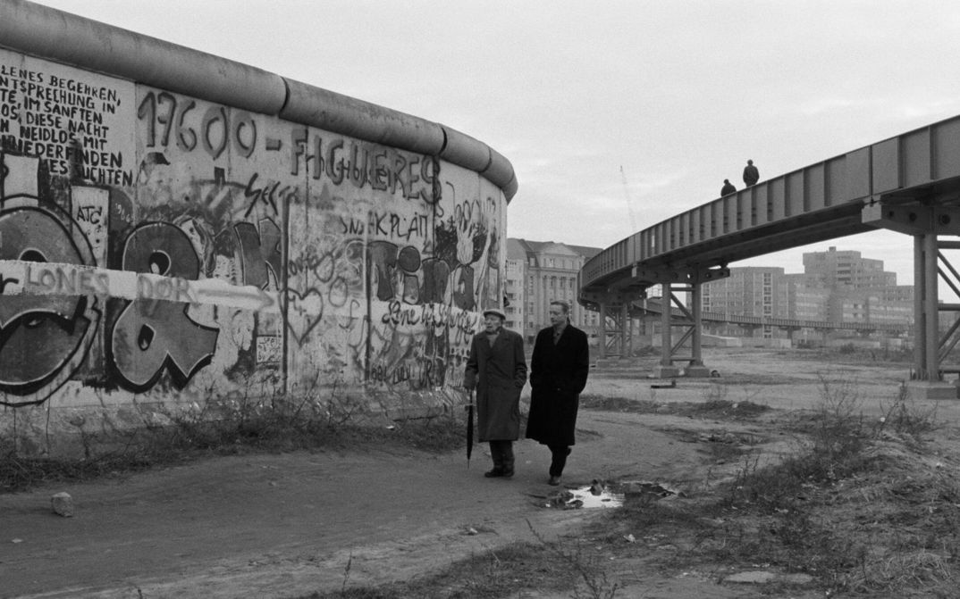 Filmstill aus DER HIMMEL ÜBER BERLIN: Zwei Männer gehen über eine Brachfläche, links von ihnen die bemalte Berliner Mauer.