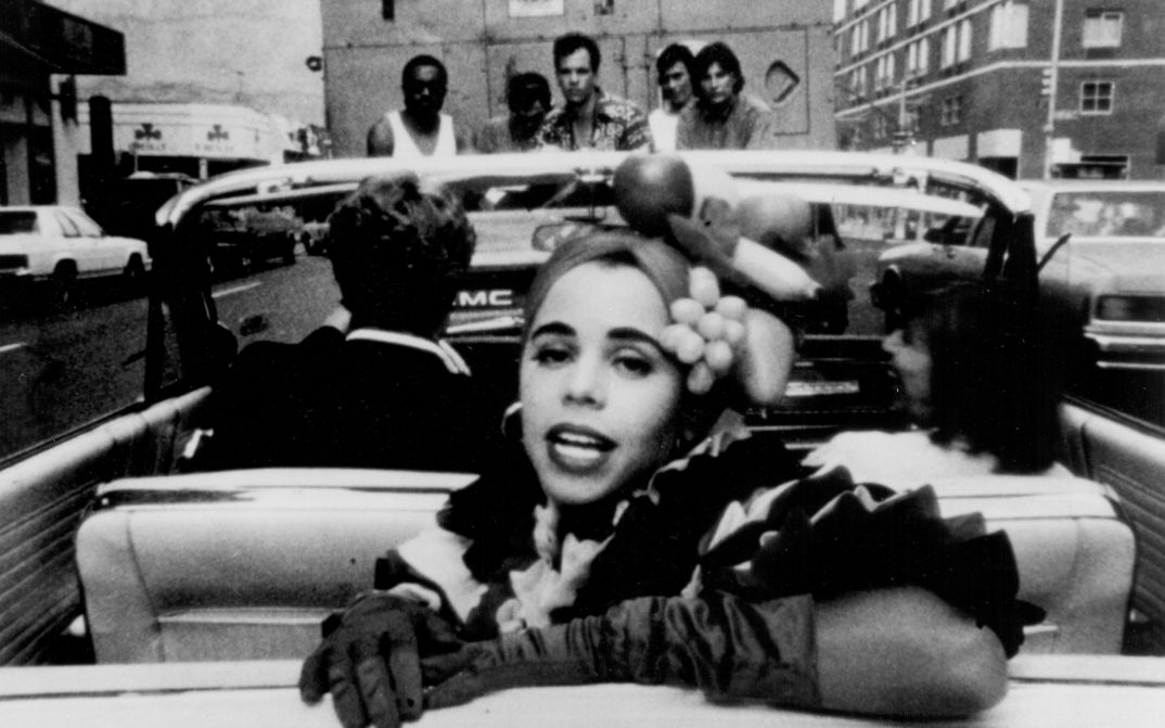Film still from PRIVILEGE: A woman turns to the viewer in the back seat of a convertible.