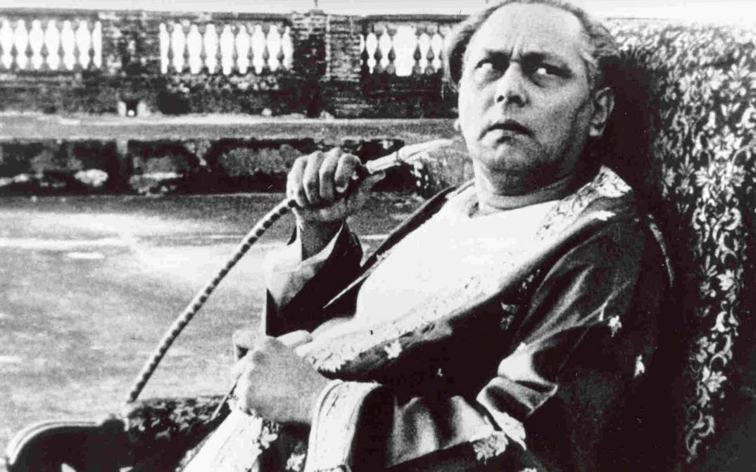 Filmstill aus JALSAGHAR: Ein älterer Mann sitz auf einem Sessel, blickt skeptisch nach oben und hält einen Shisha-Schlauch in der Hand.