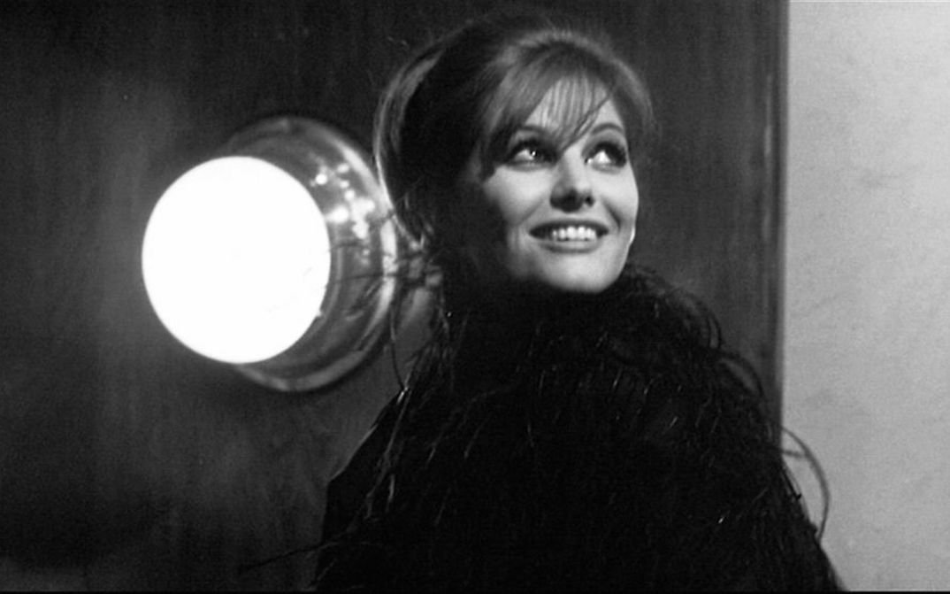 Film still from OTTO E MEZZO: A smiling young woman looks up at the sky, a bright lamp shines on the wall next to her.
