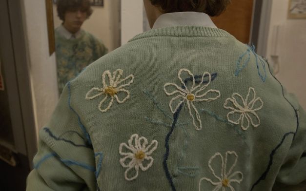 Film still from THE SWAN SONG OF FEDOR OZEROV by Yuri Semashko.The image shows the back of a man wearing a light green sweater embroidered with flowers.