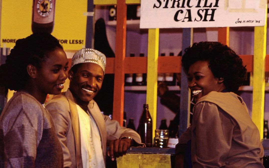 Still from the film „Der Kampf um den heiligen Baum" by Wanjiru Kinyanjui. Two women and a man standing at a bar laughing.