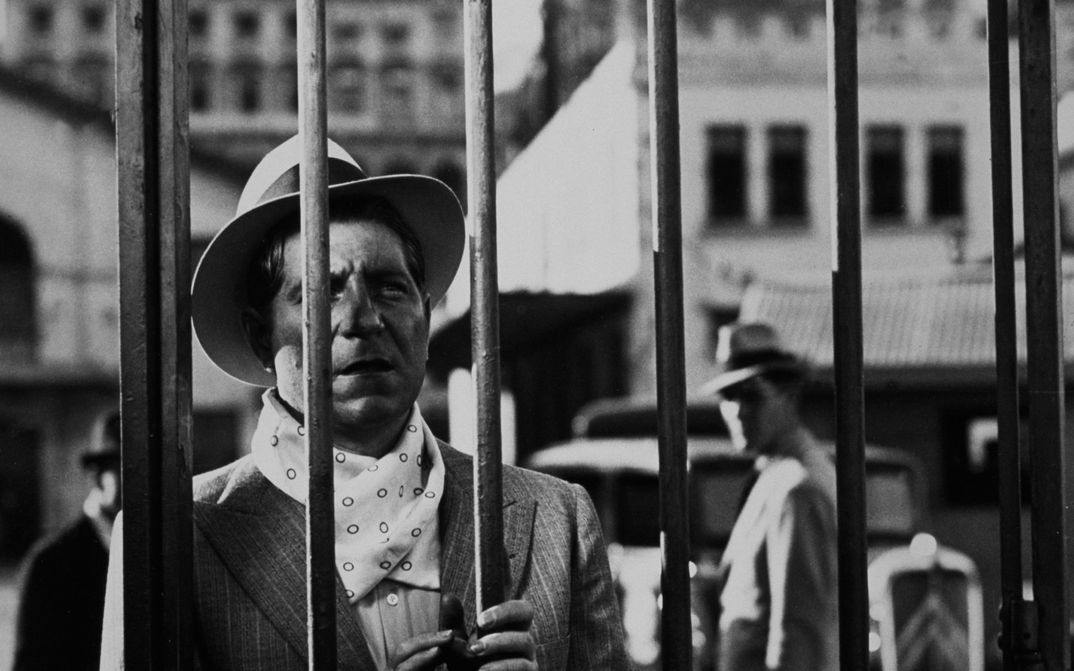 Film still from PÉPÉ LE MOKO: A man is looking through a wire fence.