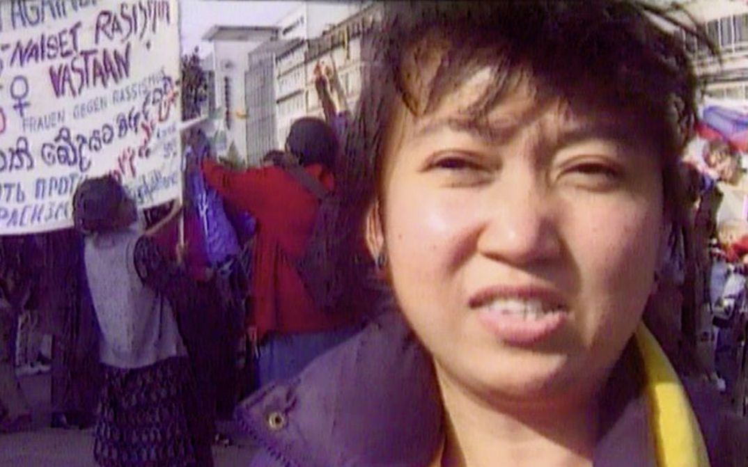 Filmstill aus „Normalität 1–10“ von Hito Steyerl. Eine dunkelhaarige Frau in Nahaufnahme, im Hintergrund eine Demo mit Menschen und Transparenten.