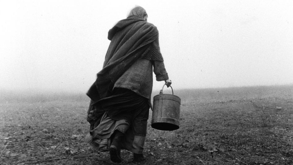 Filmstill aus DAS TURINER PFERD: Von hinten ist eine Person zu sehen, die über ein karges Feld läuft und einen Eimer trägt.