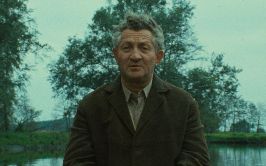 Film still from SHOAH: An elderly man in conversation, water and trees can be seen behind him.