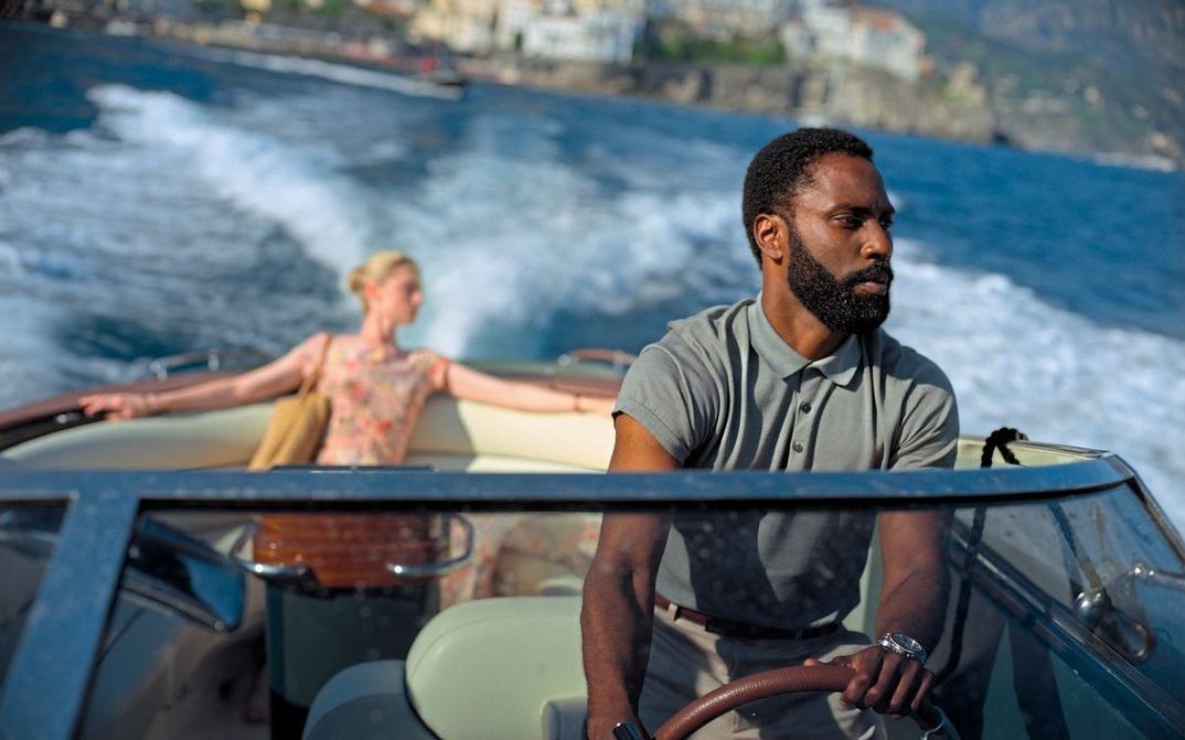 Film still from TENET: A man is at the wheel of a small motorboat, behind him sits a woman.
