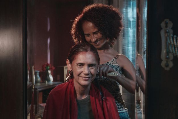 Two women stand in an interior, one seated on a chair while the other combs her hair.