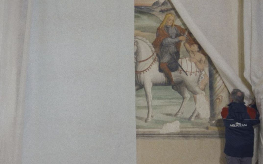 Film still from CANONE EFFIMERO by Gianluca De Serio and Massimiliano De Serio. In the picture, a man pulls back a white curtain, revealing a painting behind it.