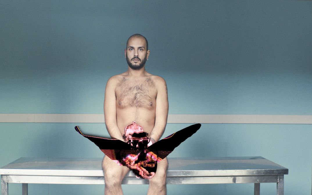 Filmstill aus EMBODIED CHORUS: Ein junger Mann sitzt nackt auf einer Bank vor einer türkis gestrichenen Wand.