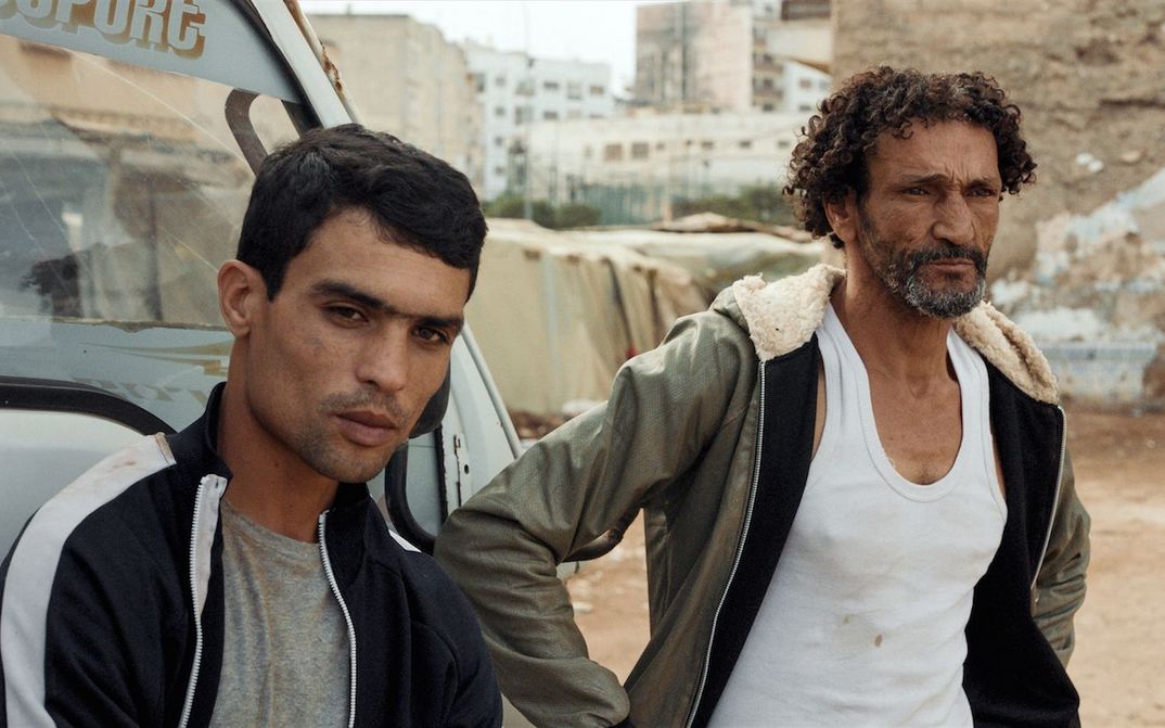 Filmstill aus HOUNDS: Zwei Männer in einfacher Kleidung stehen in einer Stadt neben einem Auto.