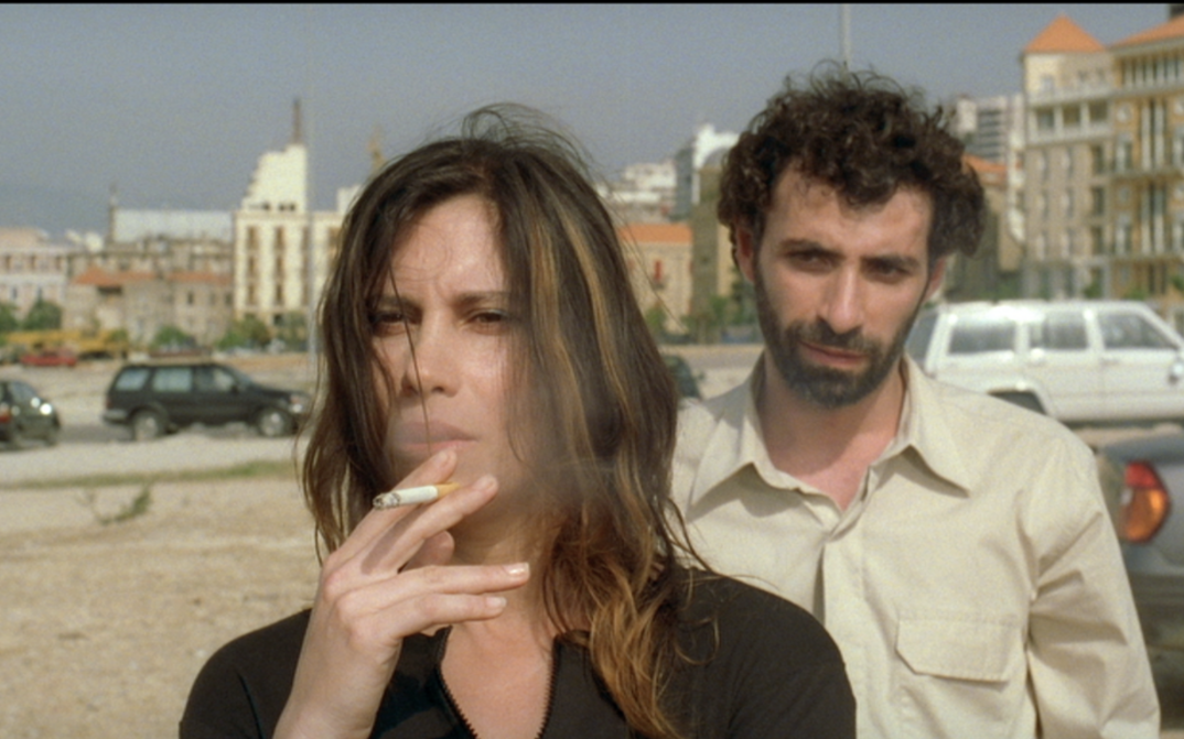 Film still from TERRA INCOGNITA. On a street stands a woman smoking, behind her stands a man.