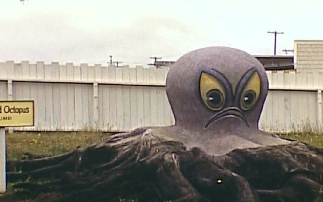 Film still from TERRA FEMME. You can see a huge octopus figure with scowling face on a meadow.