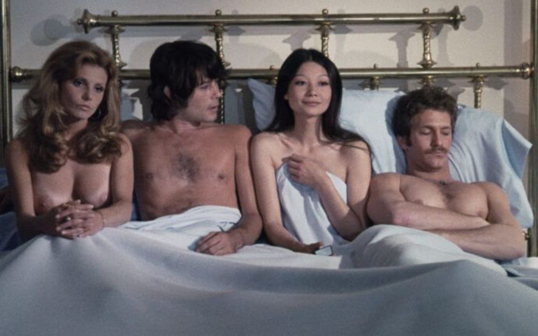 Film still from GROUP MARRIAGE: Two men and two women sit and lie unclothed in a large bed.