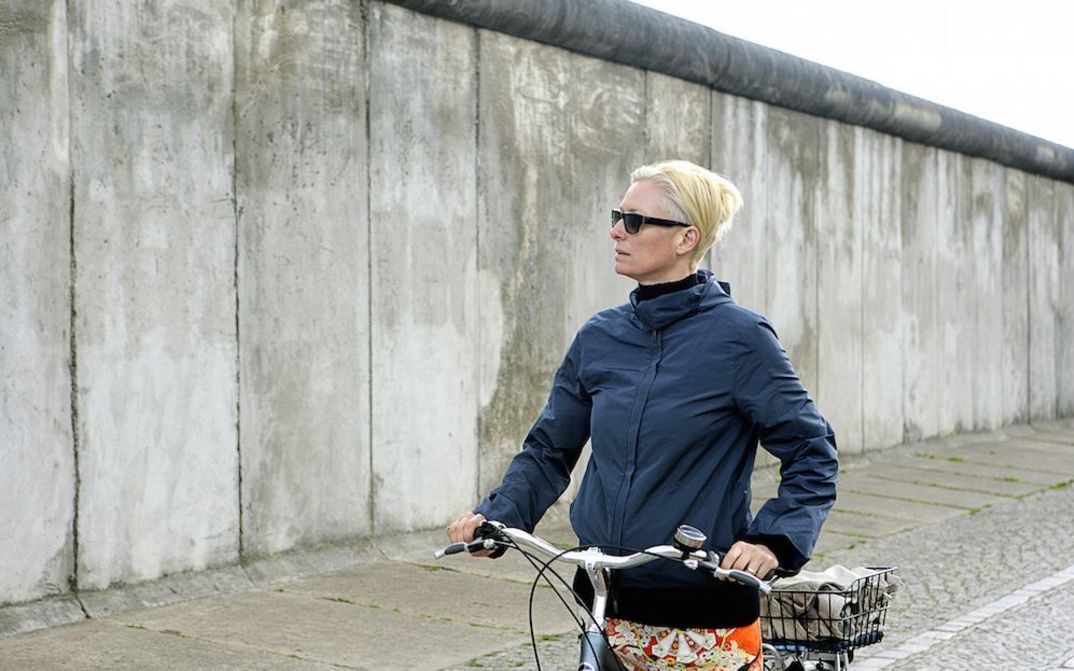 Film still from THE INVISIBLE FRAME: Actress Tilda Swinton stands with her bicycle in front of the Berlin Wall.