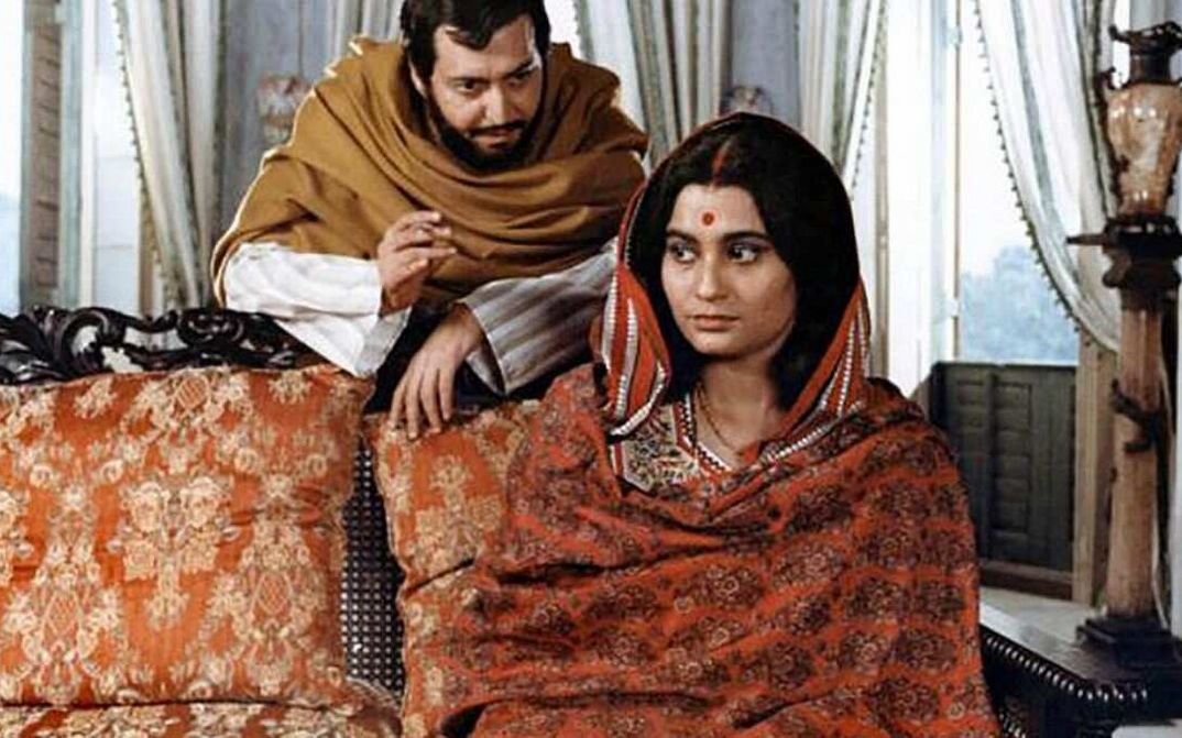 Film still from GHARE BAIRE: A woman sits on a sofa, a man stands behind her and leans towards her.
