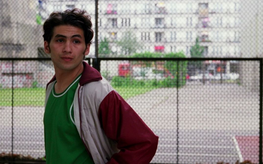 Film still from DEALER: A man in a tracksuit jacket stands in front of a fence and looks to the right. Cars, a building and green spaces can be seen in the background.