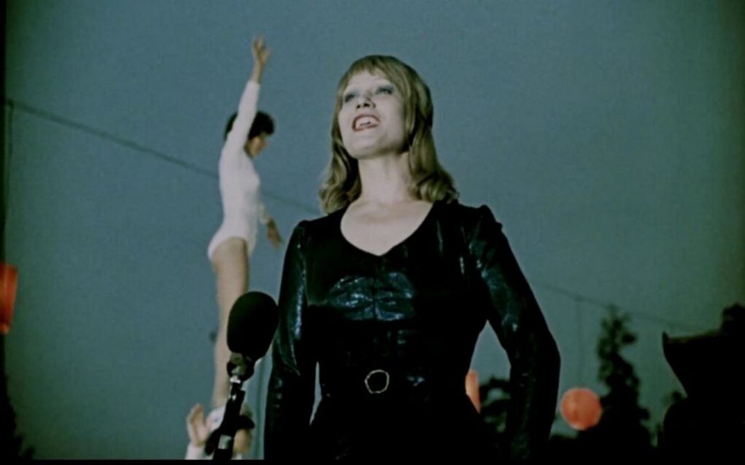 Film still from GETTING TO KNOW THE BIG, WIDE WORLD: A woman in make-up sings in front of a microphone. Behind her is a woman in a white leotard.