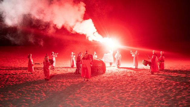 Film still from Riar Rizaldi’s film “Mirage: Eigenstate”. On a sandy, desert plateau bathed in red light, a mystical ceremony unfolds. Silhouettes form a circle, musical instruments in hand. At the center, a solitary figure rises, raising towards the sky an incandescent staff that releases swirls of smoke into the night.