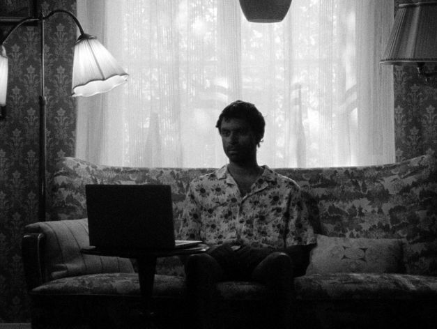 Person sitzt auf einem Sofa, vor sich ein aufgeklappter Laptop, im Hintergrund ein Fenster mit Vorhängen.