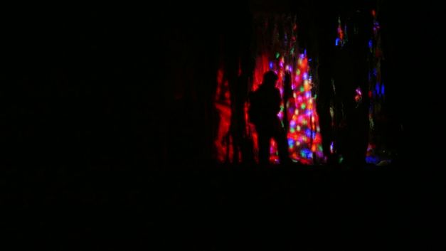 Filmstill aus dem Film „Mountain Roars“ von Chonchanok Thanatteepwong und Pobwarat Maprasob. Eine dunkle Silhouette, die sich in den Schatten einer Höhle abzeichnet, gebadet in einem Kaleidoskop aus bunten Lichtern.