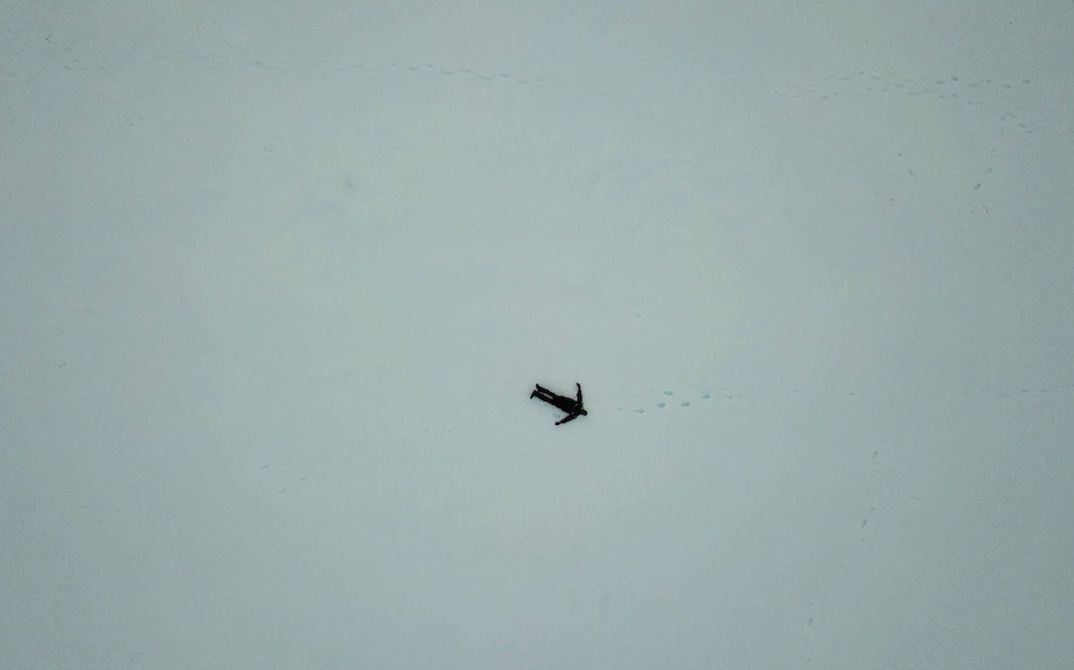 Filmstill aus Viera Čákanyovás „FREM“. Von weit oben aus der Luft fotografiert sieht man eine dunkel gekleidete, menschliche Figur in einer weiten Eiswüste liegen.