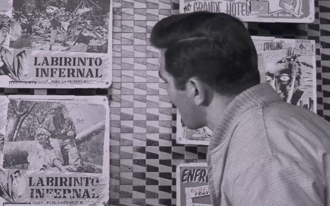 Filmstill aus BELARMINO: Ein schwarzhaariger Mann blickt weg von der Kamera auf eine Plakatwand. Auf den Plakaten steht “Labirinto Infernal”.