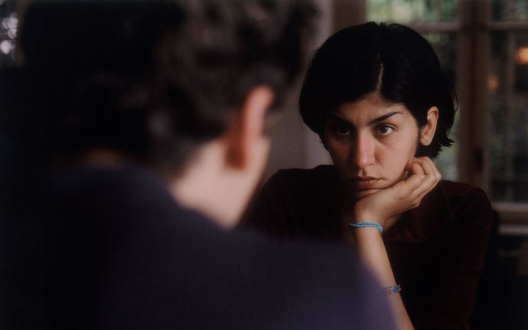 Film still from THE BEAUTIFUL DAY: Two people are sitting opposite each other. One person can only be seen from behind and the other from the front, leaning their chin on their hand. The people look at each other.