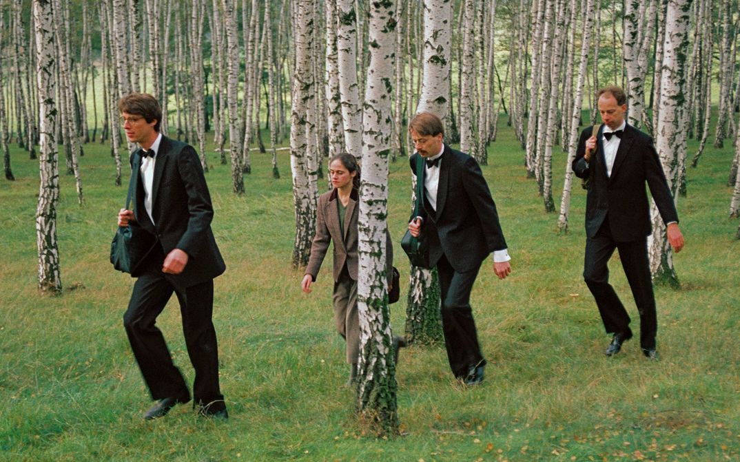 Filmstill from DIE DREI GERECHTEN KAMMACHER: 3 men in suits and bow ties and a woman in a brown suit walk in a row through a birch grove.