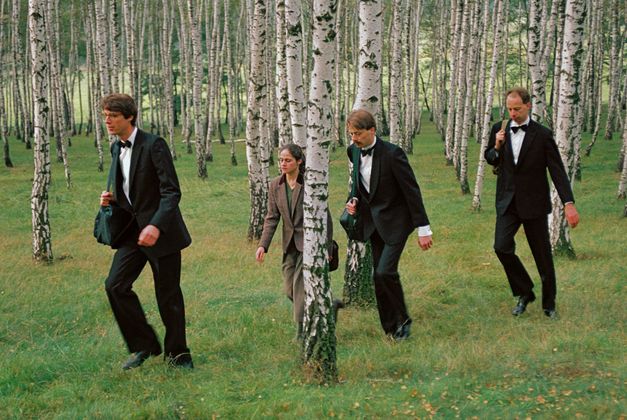 Filmstill aus DIE DREI GERECHTEN KAMMACHER: 3 Männer in Anzug und Fliege und eine Frau in braunem Anzug laufen in einer Reihe durch einen Birkenhain.