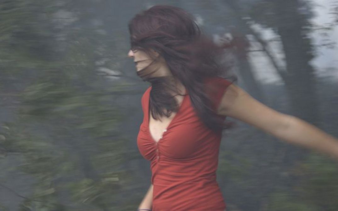 Film still from ÇA BRÛLE: A young woman with long brown hair and a red top is standing in front of a patch of woods, which you can only see out of focus. Her face is half hidden by her hair.