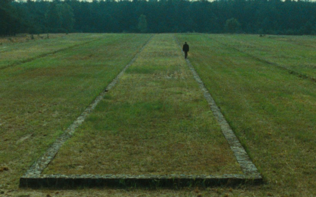 Film still from SHOAH: You can see a landscape with a meadow and forest in the background. The remains of a building can be seen in the meadow, a person is walking along it.