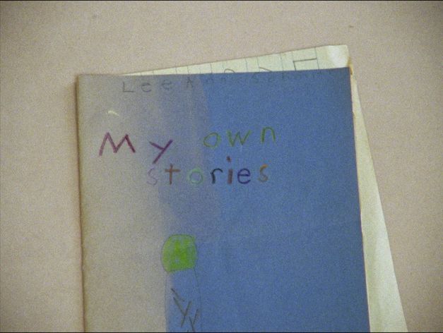 Filmstill aus EVIDENCE von Lee Anne Schmitt. Das Bild zeigt ein blaues Schulheft mit einer Kinderzeichnung und einer Aufschrift in kindlicher Handschrift auf dem Einband.