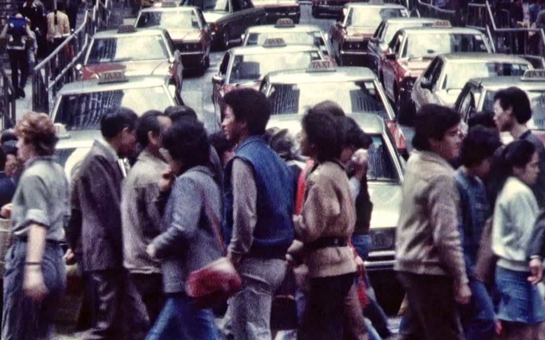 Filmstill aus HONG KONG TOPOGRAPHY. Menschen überqueren eine Straße, im Hintergrund Autos.