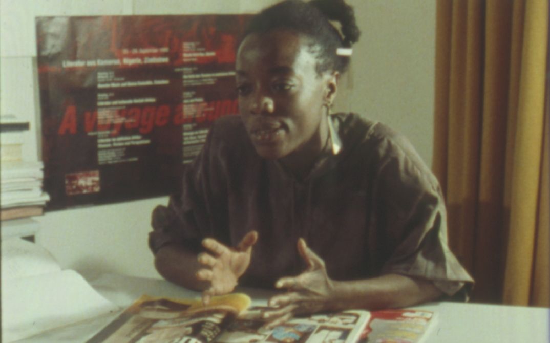 Still aus dem Film „Black in the Western World“. Eine Frau sitzt an einem Schreibtisch und spricht mit jemandem außerhalb des Bildes. Sie gestikuliert mit den Händen.