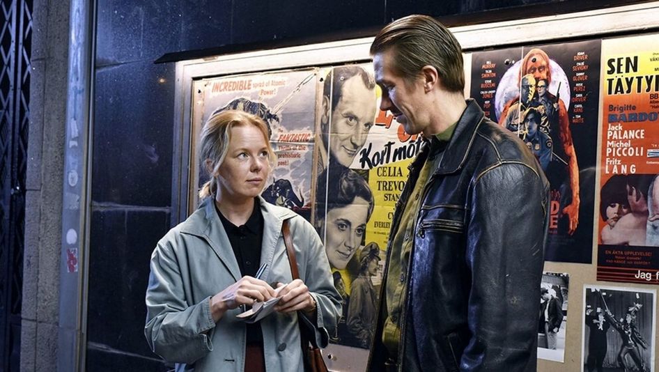 Filmstill from FALLEN LEAVES: A woman in a trench coat and a man in a leather jacket stand next to each other in front of film posters.