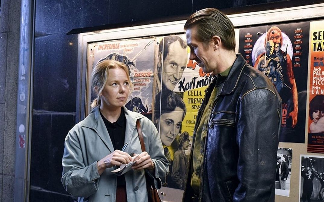 Filmstill from FALLEN LEAVES: A woman in a trench coat and a man in a leather jacket stand next to each other in front of film posters.