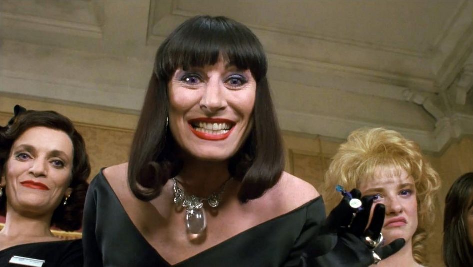 Filmstill aus THE WITCHES: Eine stark geschminkte Frau schaut mit einem bedrohlich wirkenden Lächeln auf den Boden. Hinter ihr sind zwei weitere Frauen.