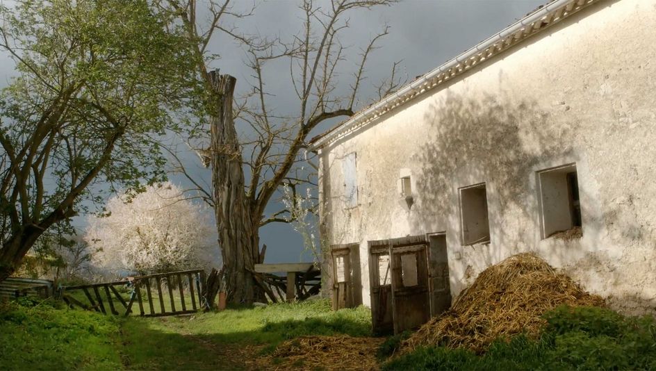 Film still from DIE FABELHAFTE WELT DES DR. CADÉOT: A whitewashed stable building with manure in front of it, you can also see trees and a fence.